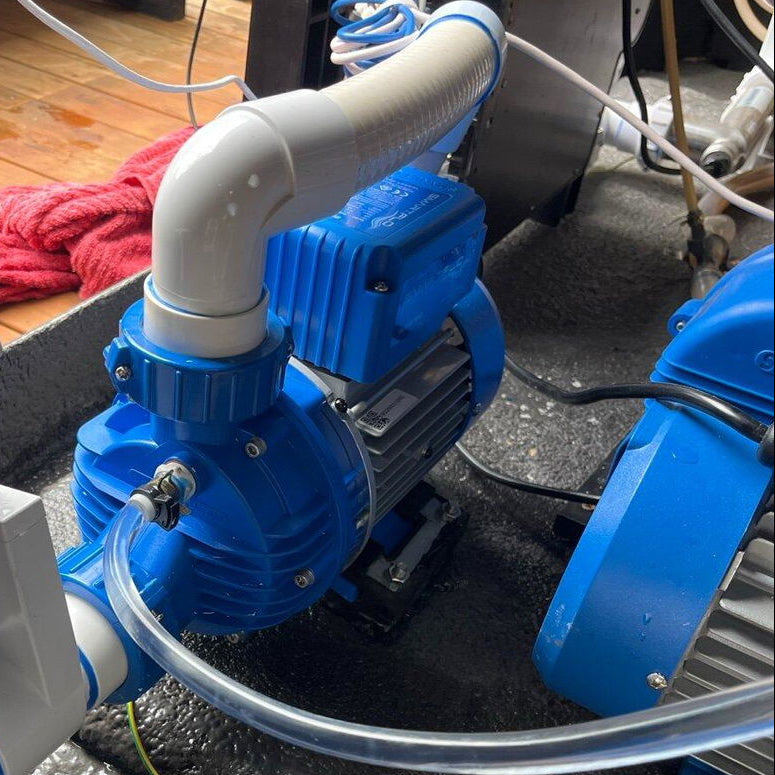 Blue and white pool pump system with clear tubing and white PVC piping on a wet concrete deck.